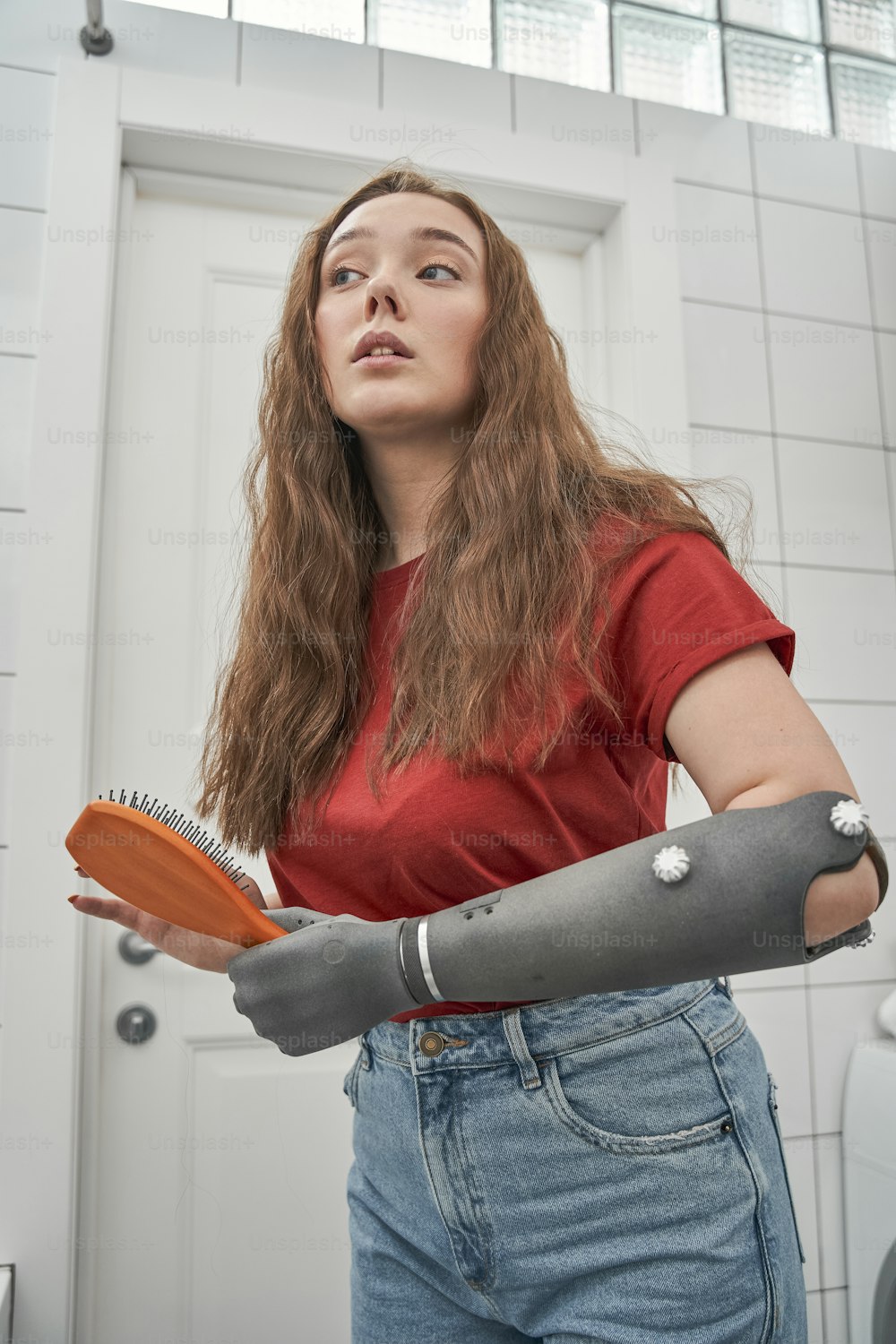 Mujer joven segura de sí misma con brazo de prótesis mirando su reflejo mientras está sentada en el espejo y cepilla el cabello pelirrojo largo y saludable. Mujer millennial haciendo rutina de belleza matutina