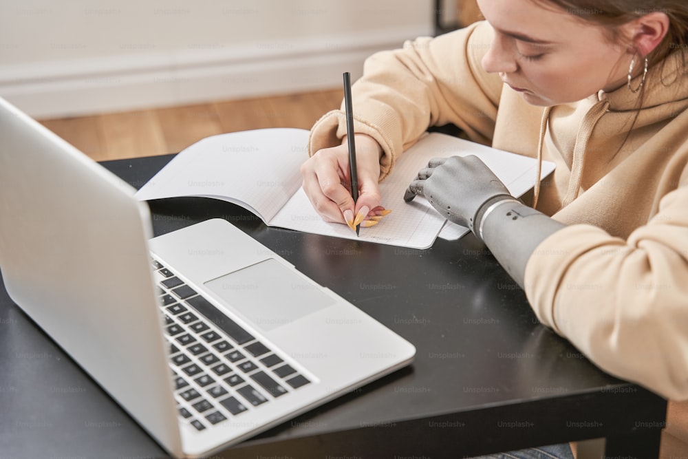 Jeune femme calme avec prothèse de bras écrivant sur le cahier tout en étant assise à la table et en faisant ses devoirs à l’ordinateur portable. Fille ayant une journée de travail à la maison