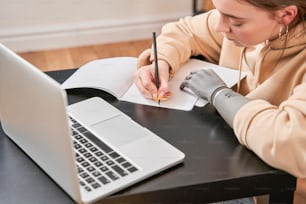 Giovane donna calma con protesi al braccio che scrive al taccuino mentre si siede al tavolo e fa i compiti al laptop. Ragazza che ha giornata lavorativa a casa