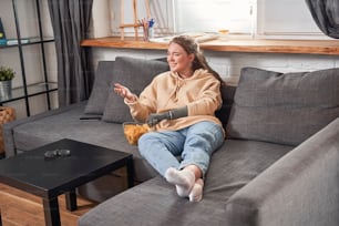 So fun. Cherry ginger woman with artificial limb holding plate with nachos chips while watching movie and laughing. Woman sitting at the sofa at home. Stock photo