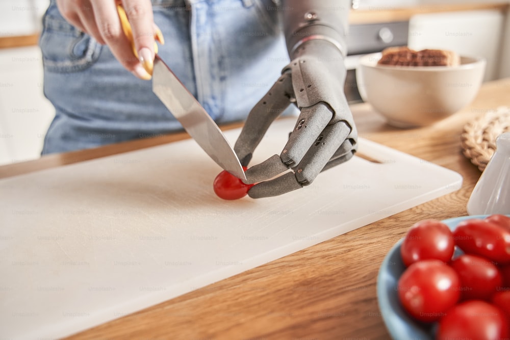 La hermosa joven pelirroja con brazo protésico está cortando ingredientes en la mesa mientras prepara la cena en la cocina. Comida saludable, cocina en casa y concepto de dieta