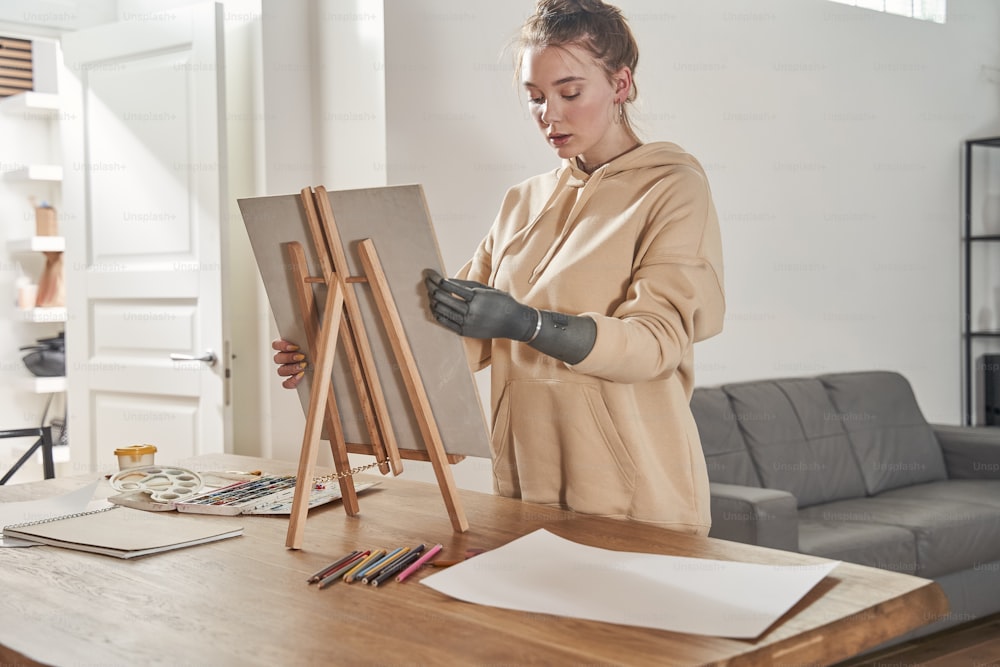 Senior artist working in concentration. Waist up portrait view of red haired woman with artificial limb holding canvas while preparing to painting pictures at easel in art studio standing at the living room