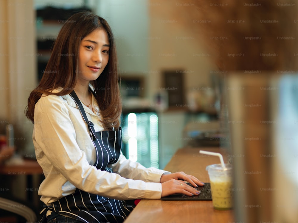Side view of female barista smiling to camera while using digital tablet on wooden bar in cafe