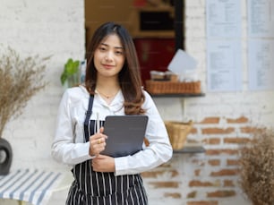 Portrait of young beautiful waitress holding digital tablet smiling to camera while standing in restaurant