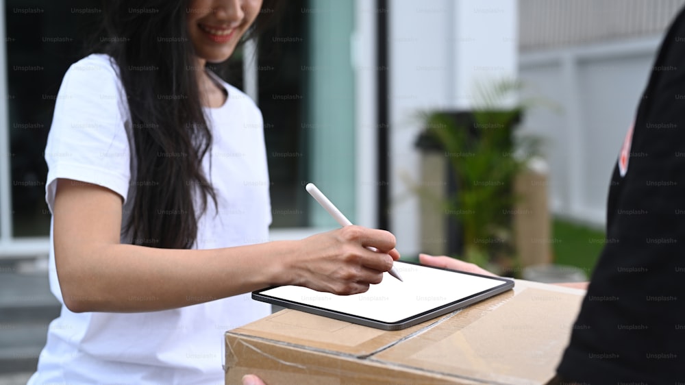 Young Asian woman customer signing on digital tablet and receiving package from delivery man at home.