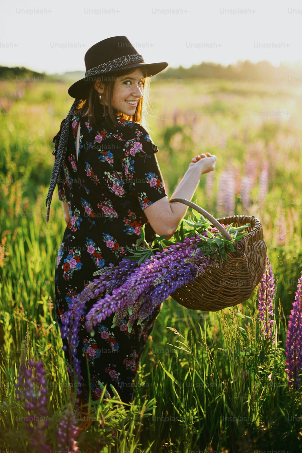 Happy beautiful woman walking in sunny lupine field, holding rustic basket with flowers. Tranquil atmospheric moment. Young female relaxing in countryside meadow at sunset.