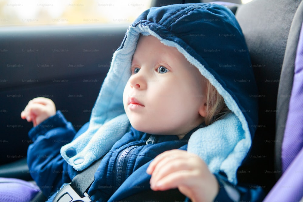 車の座席に座っているかわいい白人の赤ちゃんの幼児。自動車の車に上着を着た愛らしい子供シートベルトで固定されています。道路上の子供のケア、安全、保護。