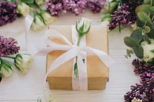 Stilvolle Geschenkbox mit Flieder- und Rosenblüten auf weißem Holzhintergrund. Happy mothers day und womens day concept. Bastelgeschenk mit Rose und Band und Blumenkomposition
