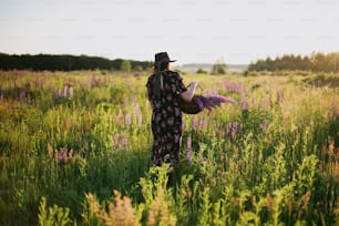 Beautiful stylish woman walking with wicker rustic basket in sunny lupine field. Tranquil atmospheric moment. Young female in vintage floral dress and hat gathering flowers in summer countryside