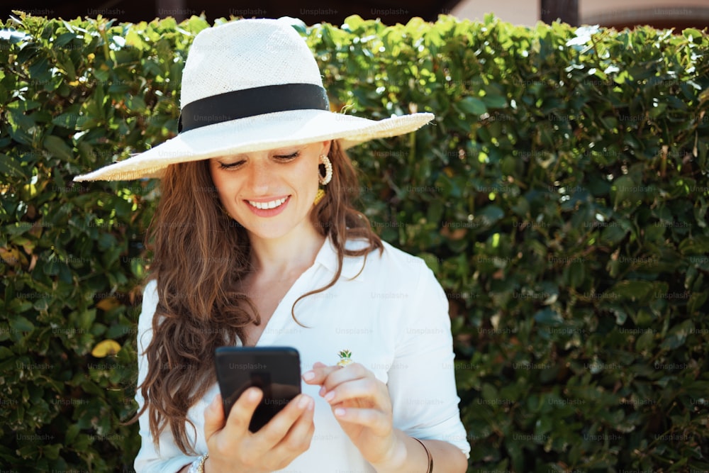 白いシャツを着た帽子をかぶった幸せなトレンディな主婦が、緑の壁の近くで屋外でスマートフォンを使用してテキストメッセージを送信します。