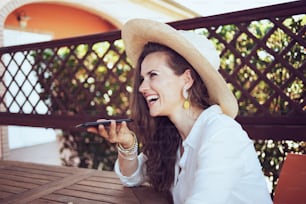 Mujer feliz de moda de 40 años en camisa blanca con sombrero sentada en la mesa usando un teléfono inteligente en la terraza del hotel de la casa de huéspedes.
