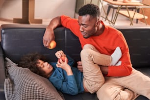 Bon appetit. Calm father and cute curly son eating apples together while spending time at the sofa at the living room. Father and son relationships concept. Stock photo