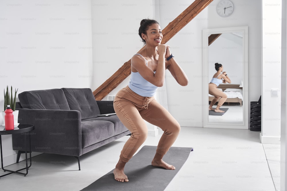 Full length view of the woman exercising at home. Sporty beautiful woman exercising to stay fit while doing squats. Fitness, workout, healthy living and diet concept. Woman doing gymnastic exercises. Woman losing weight and exercising