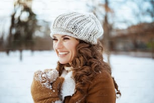 Femme souriante et élégante de 40 ans avec des mitaines dans un bonnet tricoté et un manteau en peau de mouton à l’extérieur dans le parc de la ville en hiver.