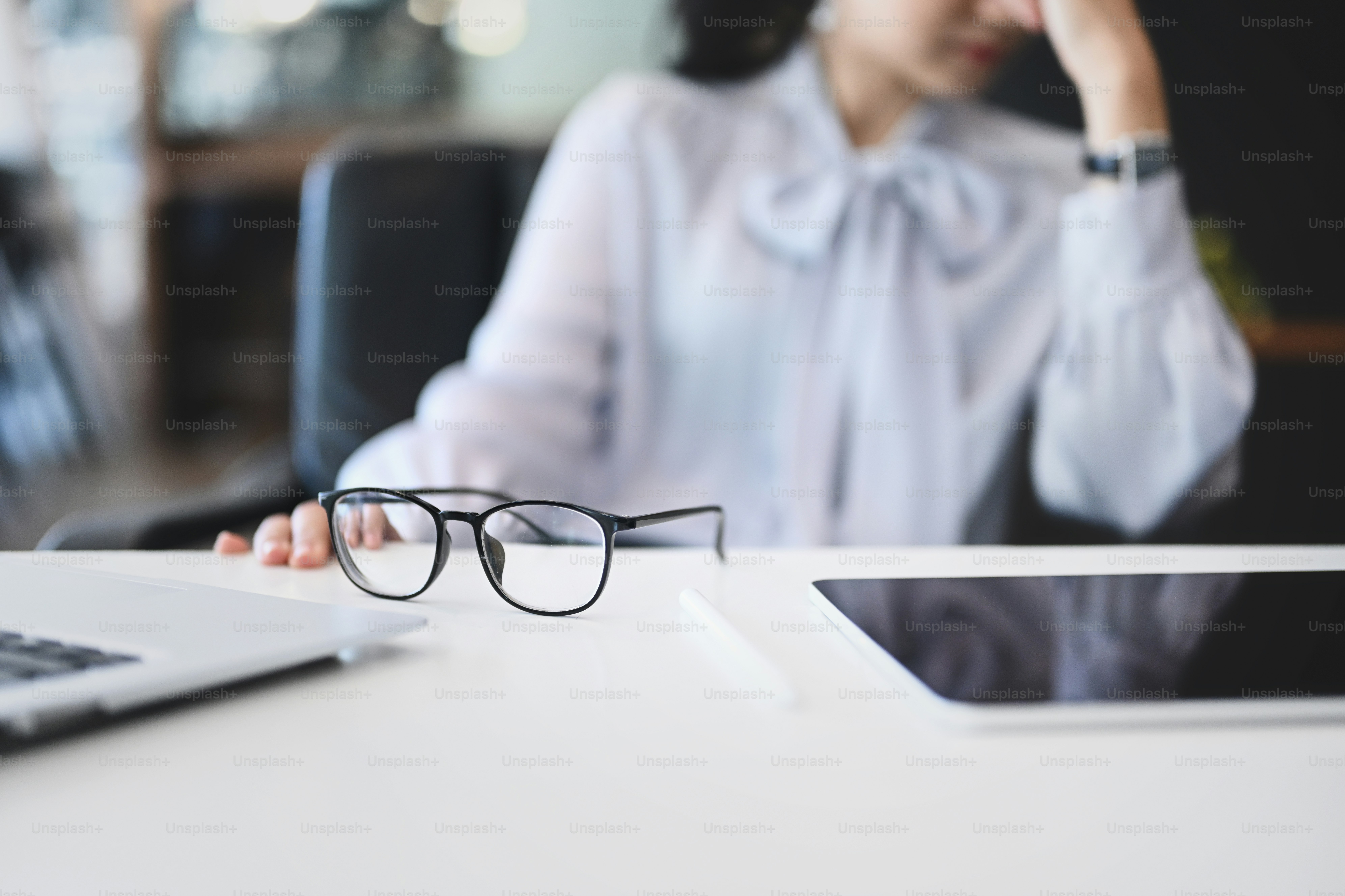 Close up view of eye glasses on office desk and businesswoman feeling exhausted in background.