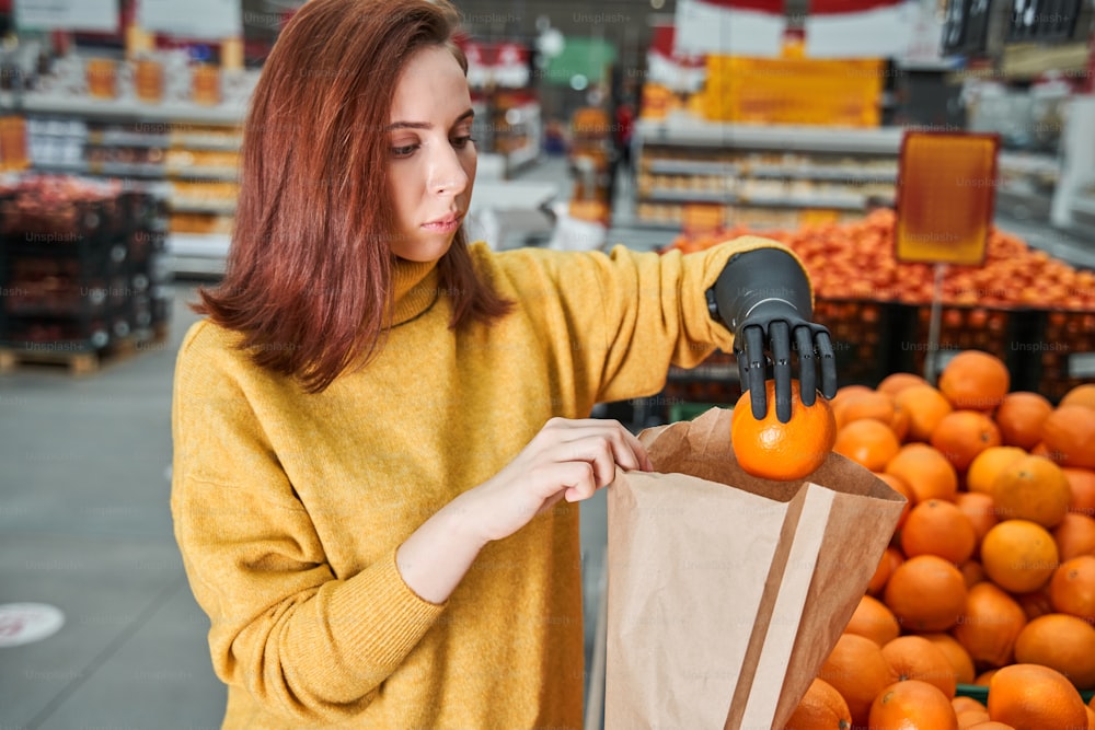 Feliz jovem encantadora mulher cliente com braço de prótese escolhendo laranjas no mercado de frutas ou supermercado. Menina colocando cuidadosamente laranjas em um saco de papel. Foto de Stock