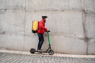 Retrato de un mensajero masculino con máscara protectora caminando con bicicleta por la calle de la ciudad, entregando comida con mochila térmica amarilla. Concepto de entrega de alimentos