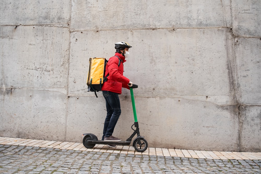보호 마스크를 쓴 남성 택배가 도시 거리에서 자전거를 타고 걷고 노란색 열 배낭으로 음식을 배달하는 초상화. 음식 배달 개념