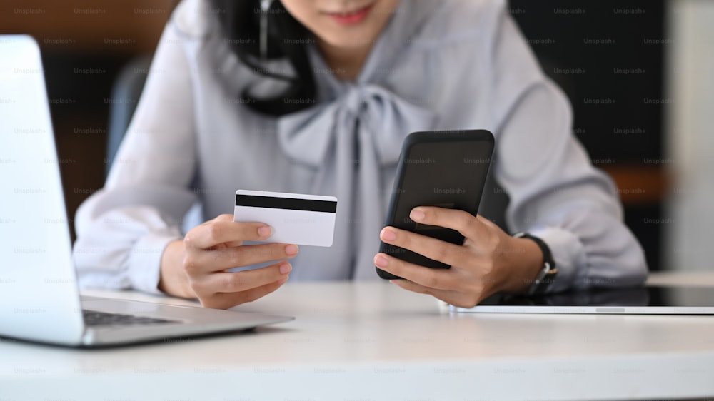 Cropped shot of young woman holding credit card and using smart phone for online shopping concept or payment transaction.