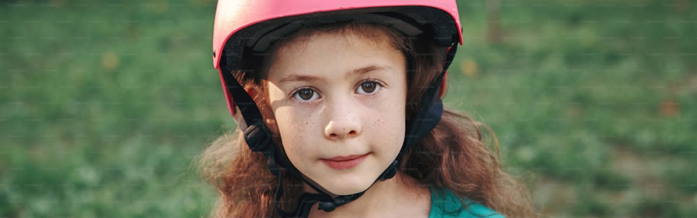 여름날 공원에서 분홍색 헬멧을 쓴 사랑스럽고 귀여운 백인 소녀의 클로즈업 초상화. 계절에 따라 야외 어린이 활동 스포츠. 건강한 어린 시절의 생활 방식. 소녀의 힘. 웹 배너 헤더.
