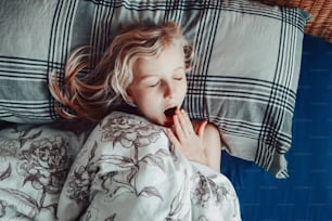 Cute adorable little child girl sleeping yawning in bed at home. Sweet dreams and bed vibes. Everyday routine early in the morning. Candid authentic home life. View from above overhead.