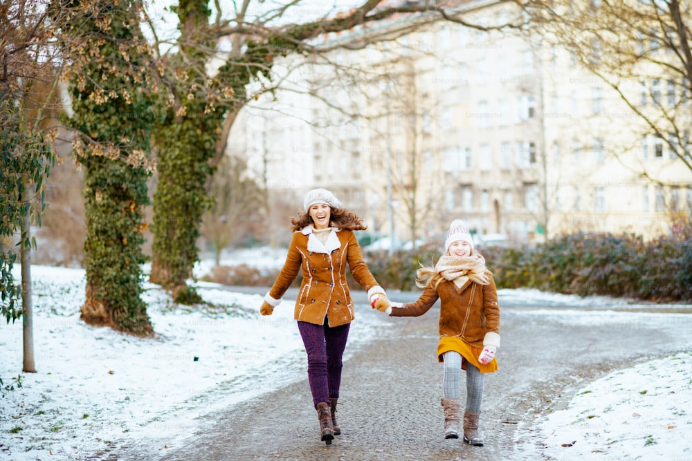 ニット帽とシープスキンコートを着た笑顔のスタイリッシュな母と娘の全身肖像画、ニット帽とシープスキンコートにミトン、冬の都市公園を屋外で散歩。