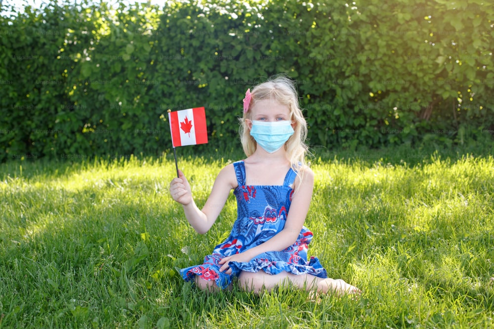 얼굴 보호 마스크를 쓴 백인 소녀가 야외에서 캐나다 국기를 흔들고 있다. 코로나바이러스 전염병 동안 캐나다의 날 휴일을 축하하는 공원의 잔디밭에 위생 마스크를 쓴 아이 아이.