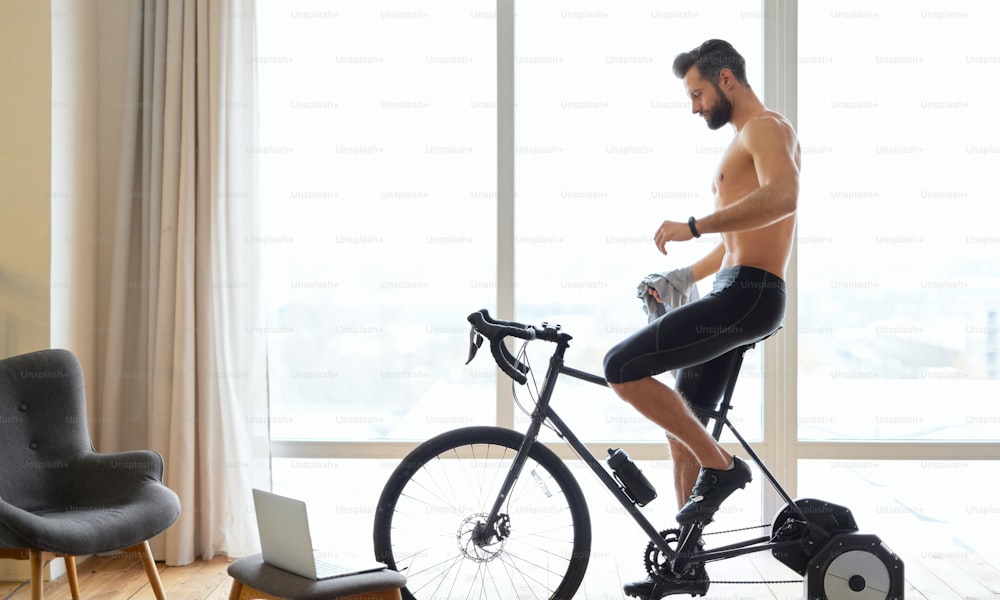 Muscular young man riding stationary exercise bike in room with laptop and large windows