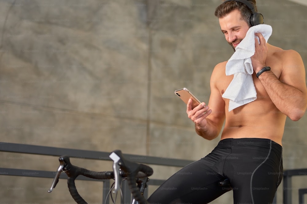 Hombre atlético alegre que lee el mensaje en el teléfono inteligente y sonríe mientras está sentado en una bicicleta estática y se limpia la cara con una toalla