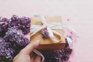 Giving gift, hand holding gift box with ribbon and lilac flowers on pink paper. Happy mothers day and valentine's day concept. Purple lilac flowers bouquet with craft present box.