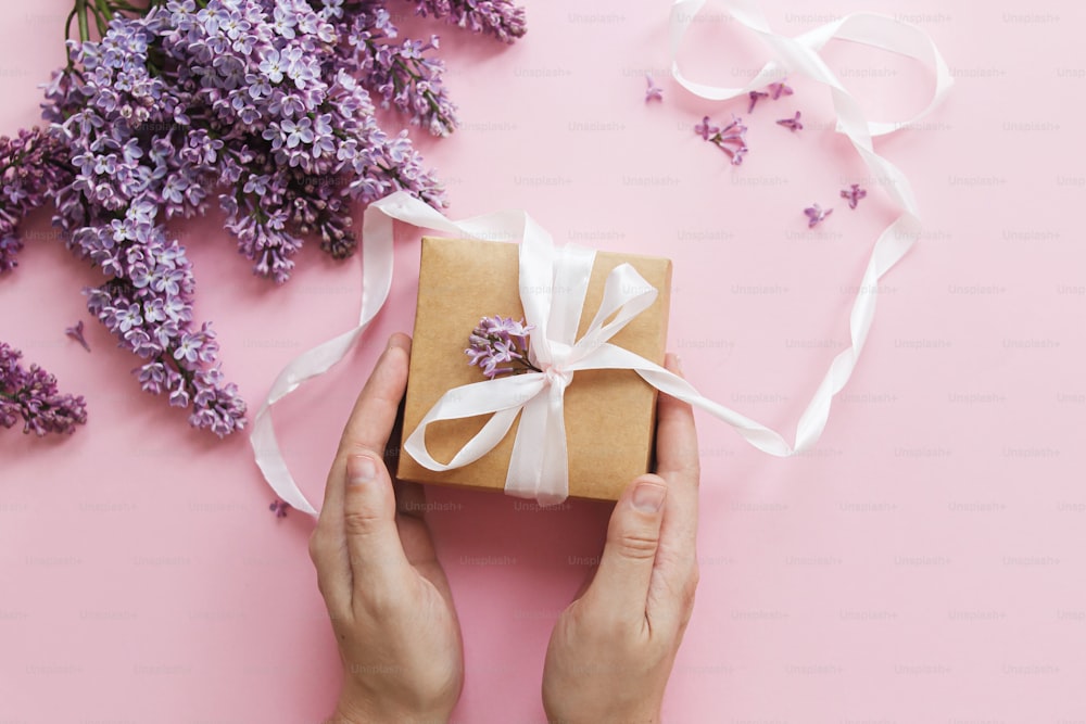 Happy mothers day and valentine's day concept. Hands holding gift box with ribbon and lilac flowers on pink paper flat lay. Purple lilac flowers bouquet with craft present box. Giving gift