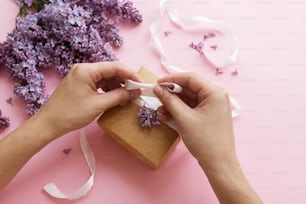Manos envolviendo caja de regalo con cinta y flores lilas en papel rosa, vista superior. Feliz día de las madres y concepto del día de San Valentín. Ramo de flores lilas moradas con caja de regalo artesanal