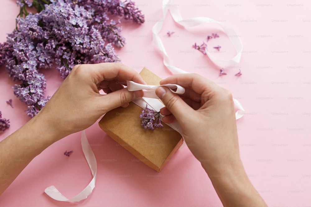 Hände wickeln Geschenkbox mit Band und lila Blumen auf rosa Papier, Draufsicht. Alles Gute zum Muttertag und zum Valentinstag. Lila lila Blumenstrauß mit Bastelgeschenkbox