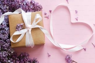 Feliz día de San Valentín y concepto del día de la madre. Flores lilas, caja de regalo y cinta de corazón sobre papel rosa. Elegante tarjeta de felicitación floral. Ramo de lilas púrpura con elegante regalo artesanal