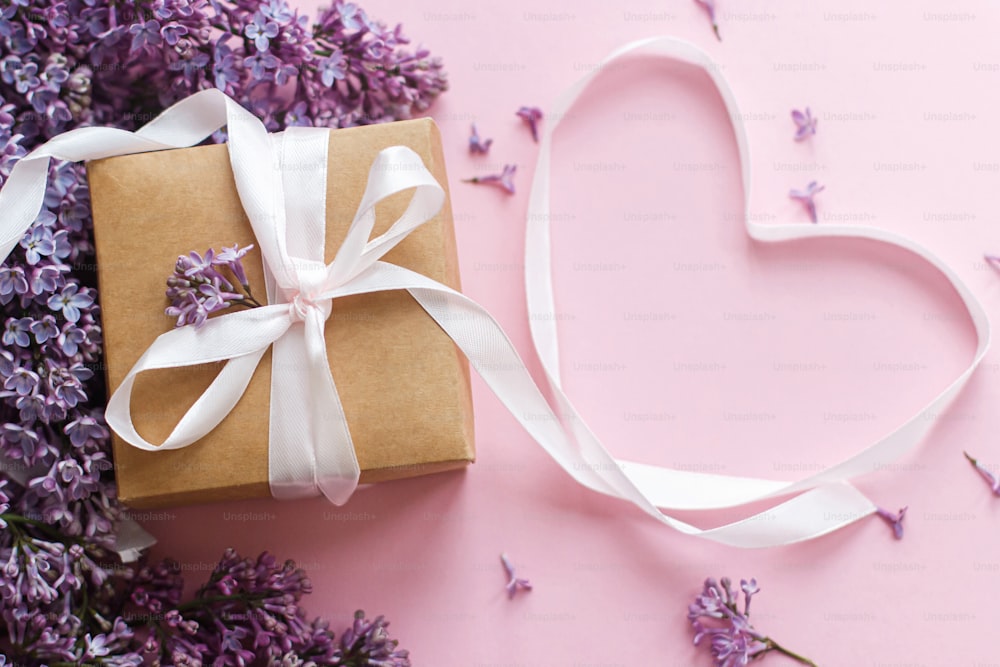 Alles Gute zum Valentinstag und Muttertagskonzept. Fliederblüten, Geschenkbox und Herzband auf rosa Papier. Stilvolle Blumengrußkarte. Lila Fliederstrauß mit stilvollem Bastelgeschenk