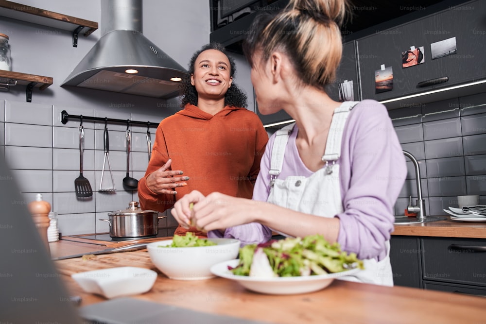 Waist up portrait of the multiracial woman chatting with her bestie while preparing salad at the table. Girl cooking with her friend at the kitchen. Her best friend telling something and laughing
