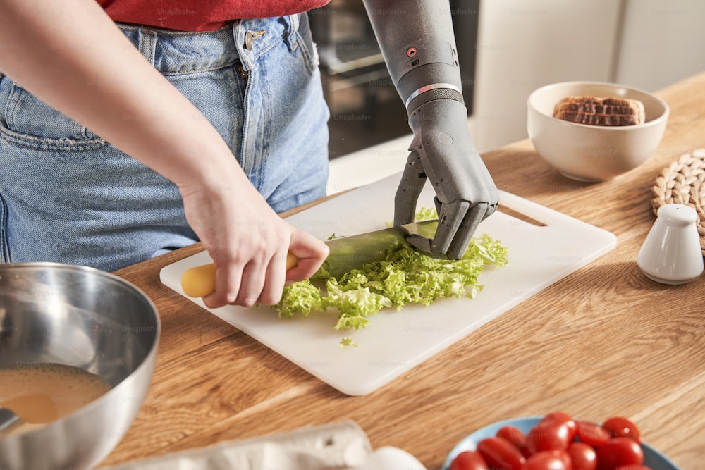 Close up view of the ginger caucasian woman with artificial limb chopping salad with a knife at the plastic plank at the table while cooking at the kitchen. Disabled people concept