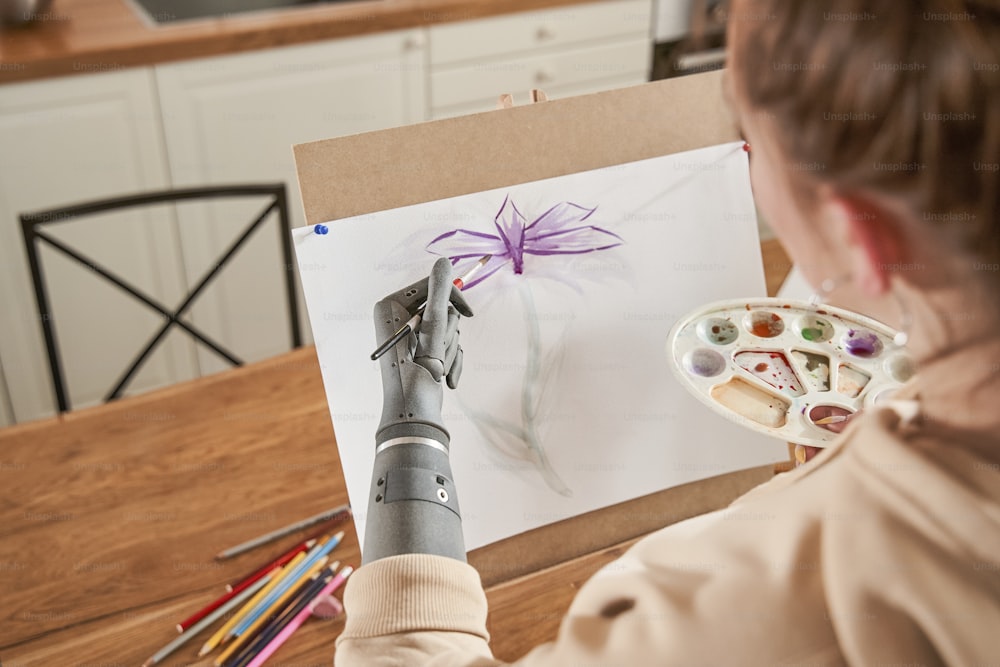 義肢を持つゴージャスで陽気な女性アーティストがワークショップのイーゼルの前に立って絵を描いている接写。水彩絵の具を使って静物画を描く少女