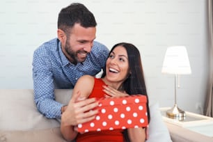 Man surprising his girlfriend with a gift on the couch at home on Valentine's Day. Beautiful young couple is celebrating at home. Man is giving his wife a gift box