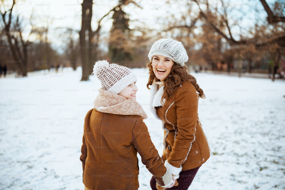 ニット帽とシープスキンコート、ミトン、ニット帽、シープスキンコートを着たエレガントな母と子が、冬の都市公園の屋外で微笑んでいます。