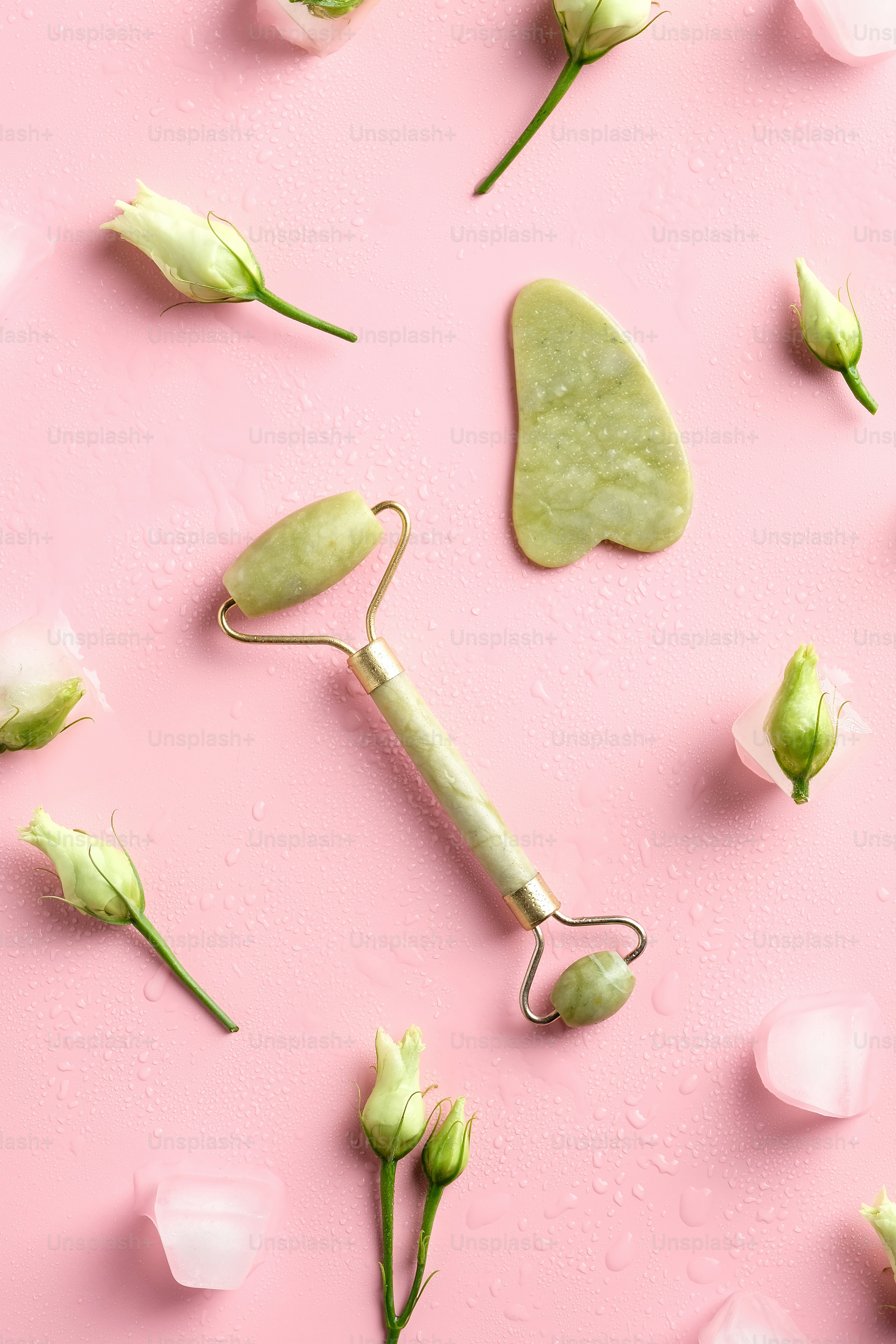 Gua sha, jade stone face roller, ice cubes, spring flowers on pink background. Facial skin treatment concept. Flat lay, top view.
