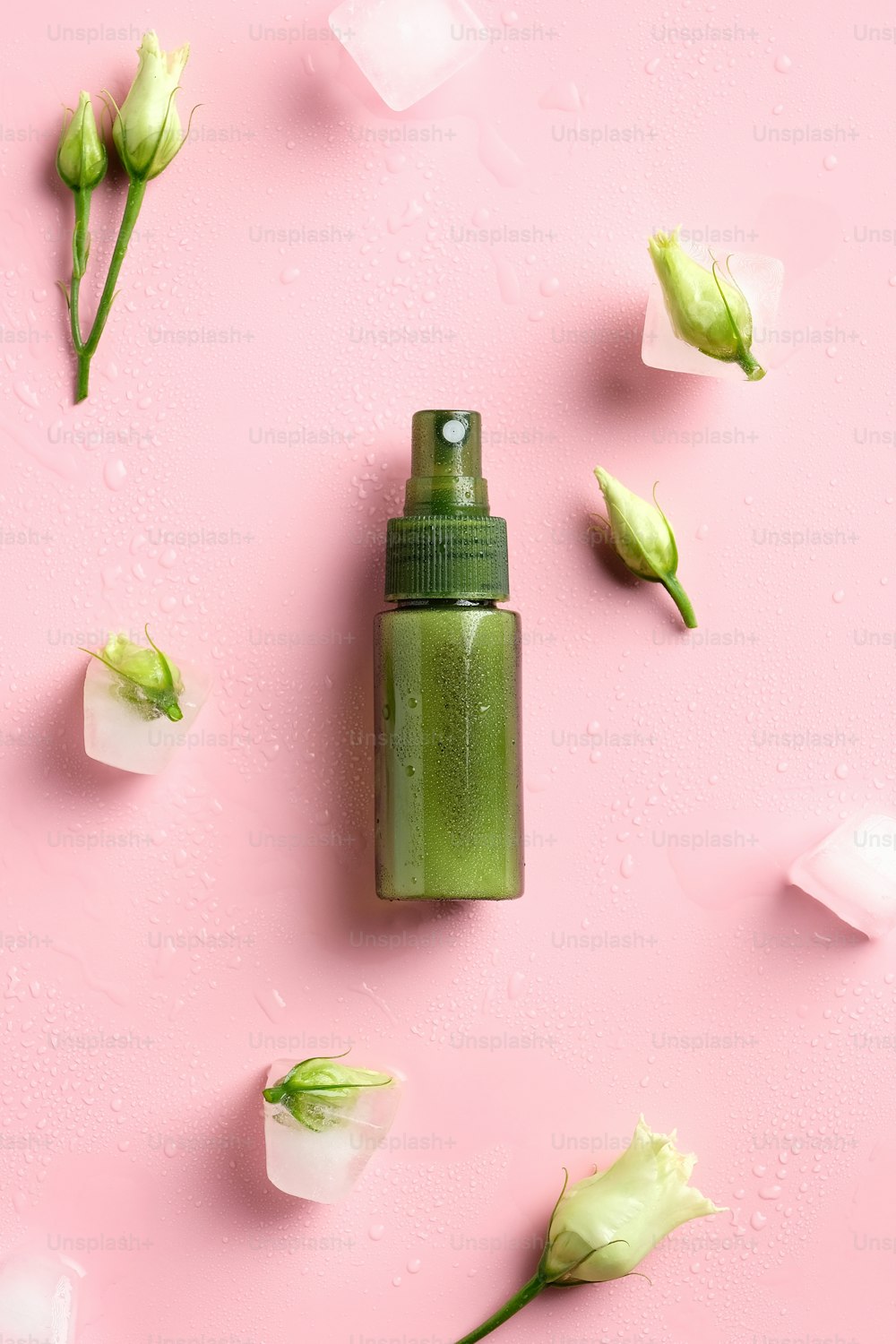 Green spray bottle, ice cubes and spring flowers on pink background. Flat lay, top view. Natural cosmetics concept.
