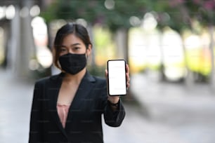 Asian businesswoman in protective mask holding and showing mobile phone with blank screen.