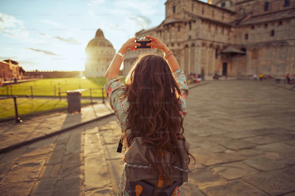 Seen from behind young woman in floral dress with vintage camera and backpack near Duomo di Pisa.
