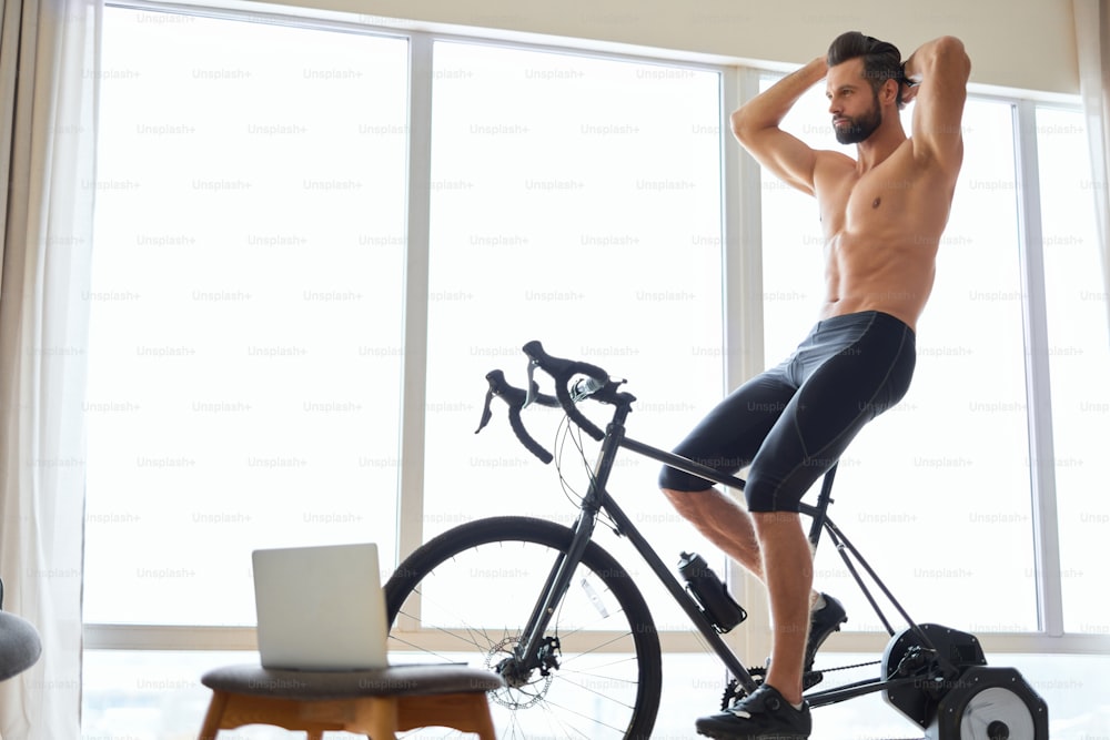 Caballero musculoso guapo con cuerpo perfecto montando bicicleta estática en la habitación con computadora portátil y ventanas grandes
