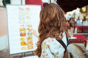 Seen from behind woman in floral dress with backpack looking at the menu in a cafe.