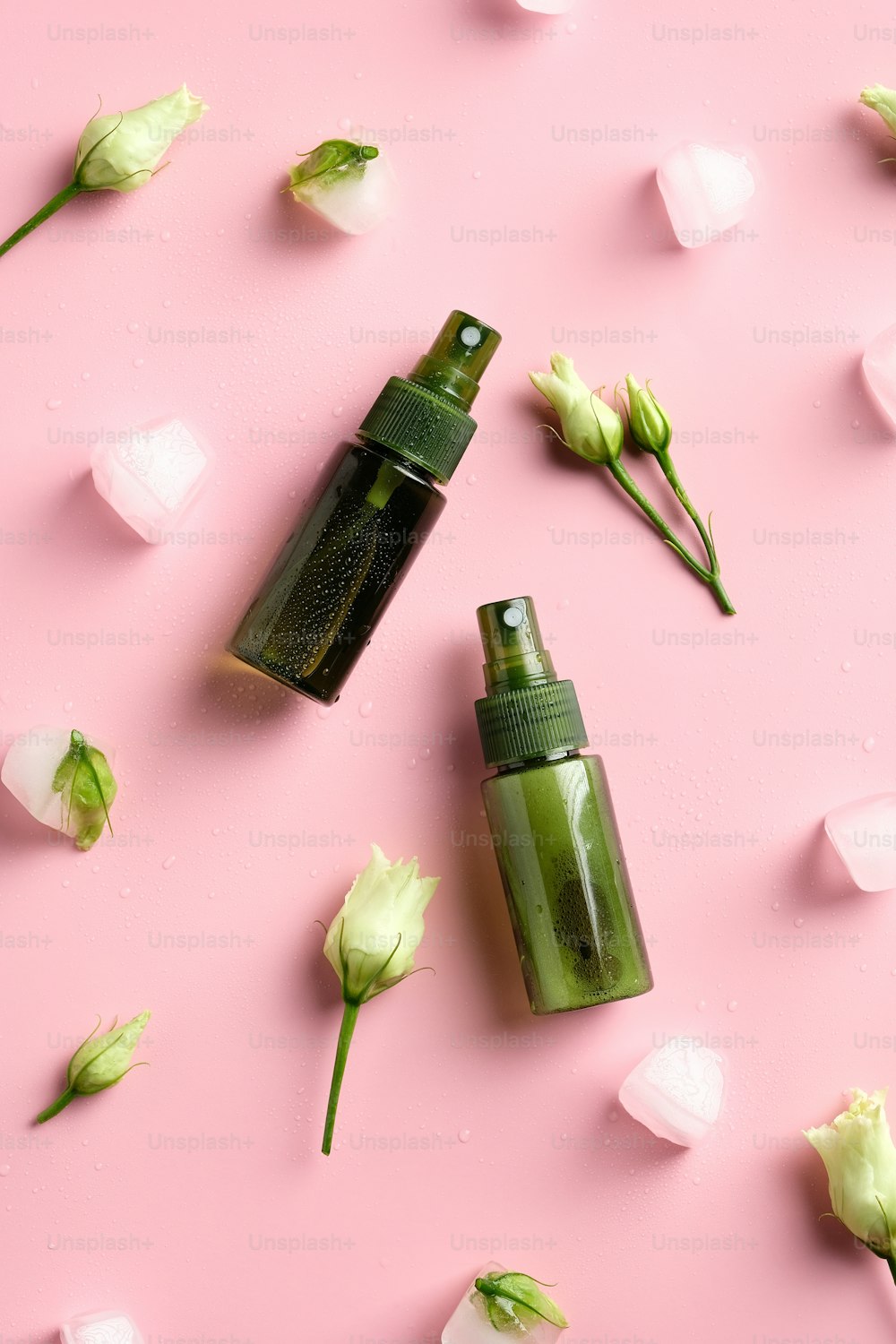 Green spray bottles with ice cubes and flowers on pink background. Natural organic skin care beauty products. Flat lay, top view.