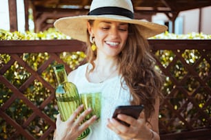 Femme souriante et élégante de 40 ans en chemise blanche avec bouteille de limonade, verre et chapeau envoyant un message texte à l’aide d’un smartphone dans le patio.