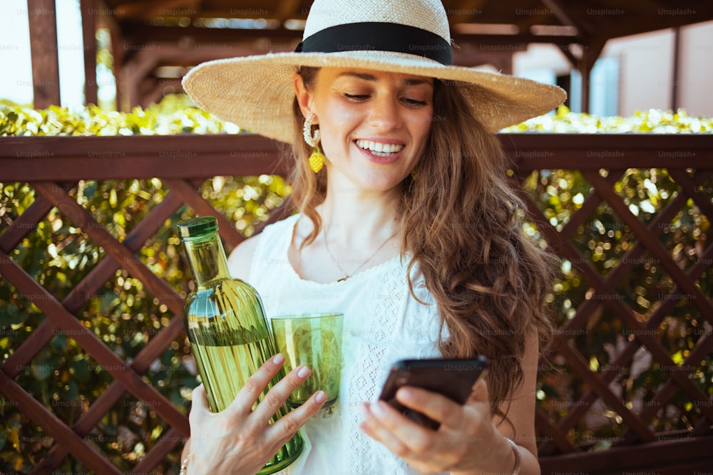 Femme souriante et élégante de 40 ans en chemise blanche avec bouteille de limonade, verre et chapeau envoyant un message texte à l’aide d’un smartphone dans le patio.