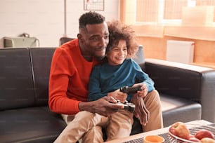 Family leisure time. Happy multiracial unshaven father and his curly stylish son playing video games on console with joysticks, while enjoying weekend at home with good emotions. Stock photo